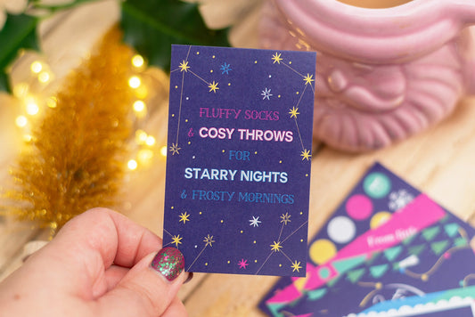 Hand holding a purple card with starry night design and text, with festive decorations in the background.