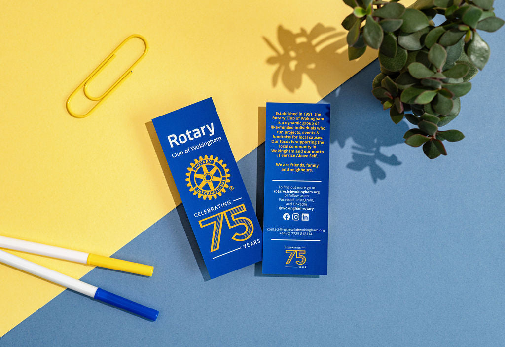 Two blue Rotary Club bookmarks on a yellow and blue background with a paperclip and plant.