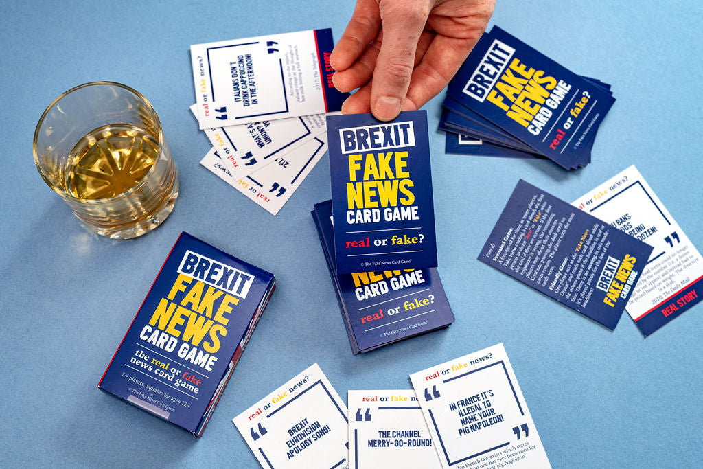 Brexit Fake News Card Game packaging and cards on a blue surface with a glass of yellow liquid.