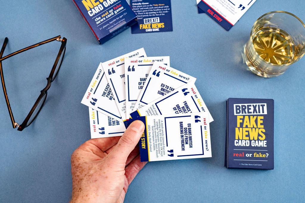 Hand holding Brexit Fake News Card Game cards on a blue surface with game box and glass of wine.