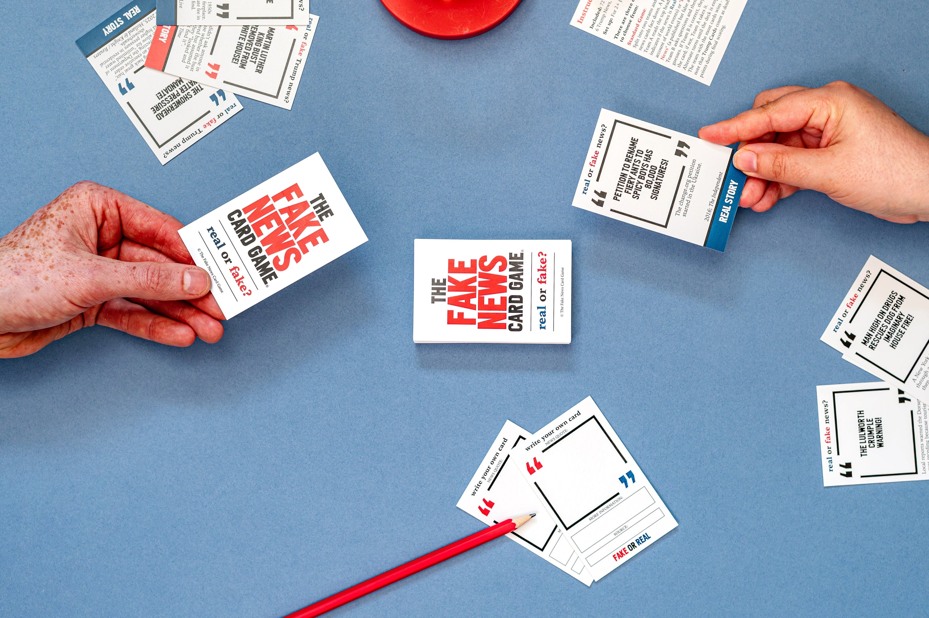 Hands playing with 'The Fake News Card Game' on a blue surface.