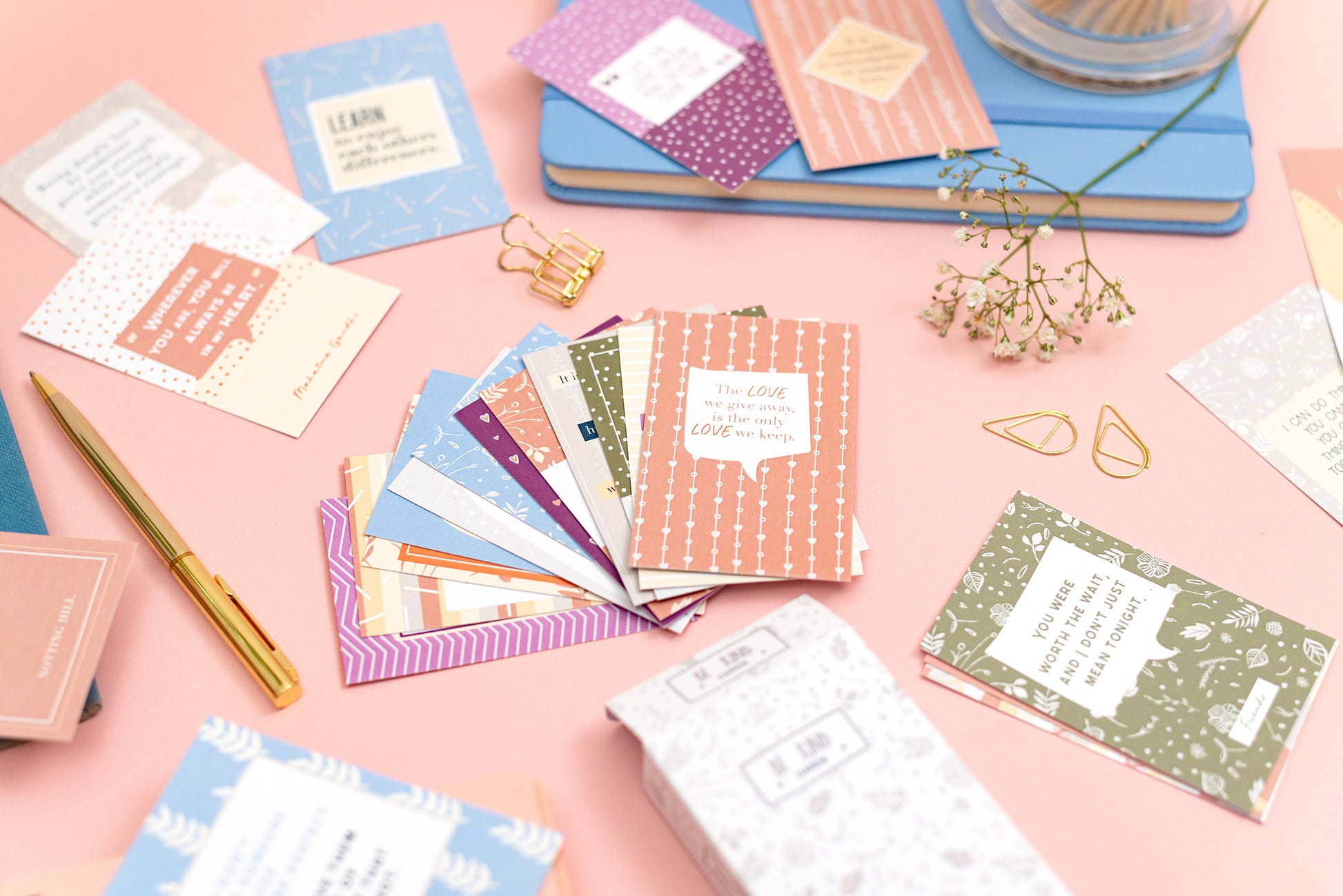 Selection of love quote cards on a pink desk surrounded by blue and gold stationery, with hedgerow flowers.