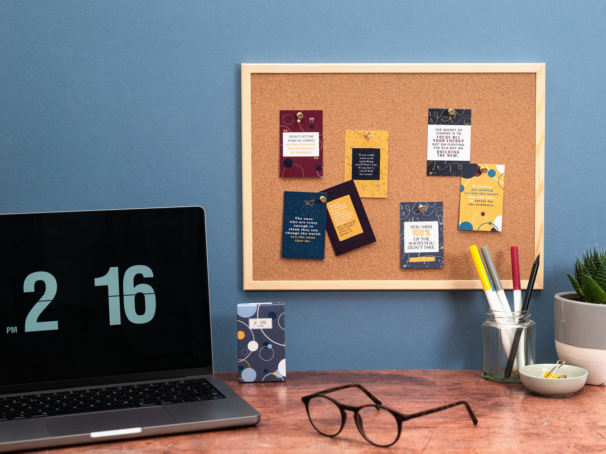 Laptop on a desk with a cork bulletin board, glasses, and stationery items against a blue wall.