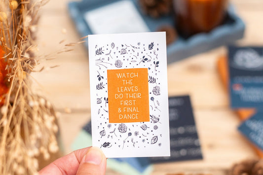 Hand holding a card with autumn-themed text and design, blurred background with fall decor.