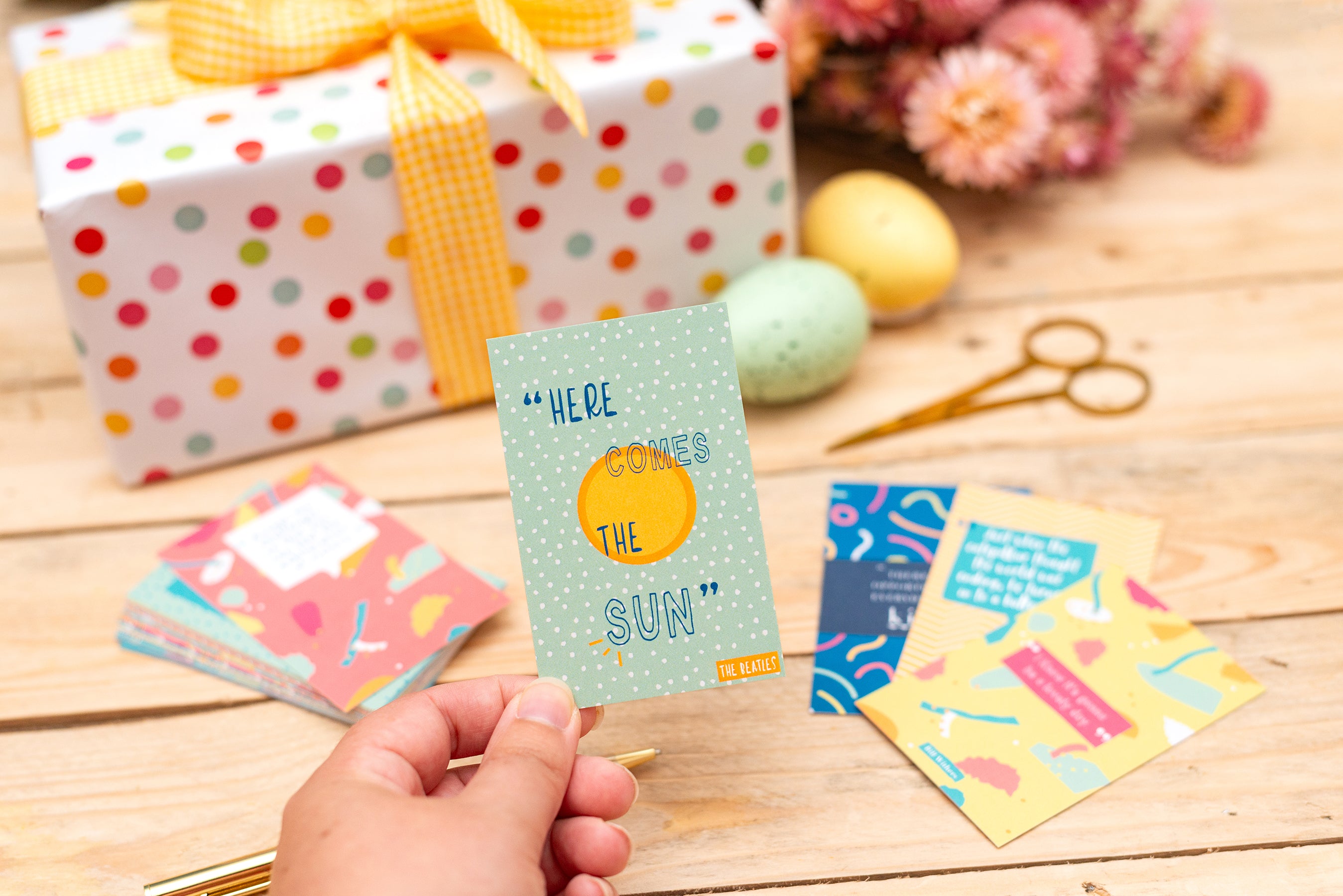 Hand holding a card with 'Here comes the sun' text, surrounded by colorful cards and a gift box on a wooden surface.