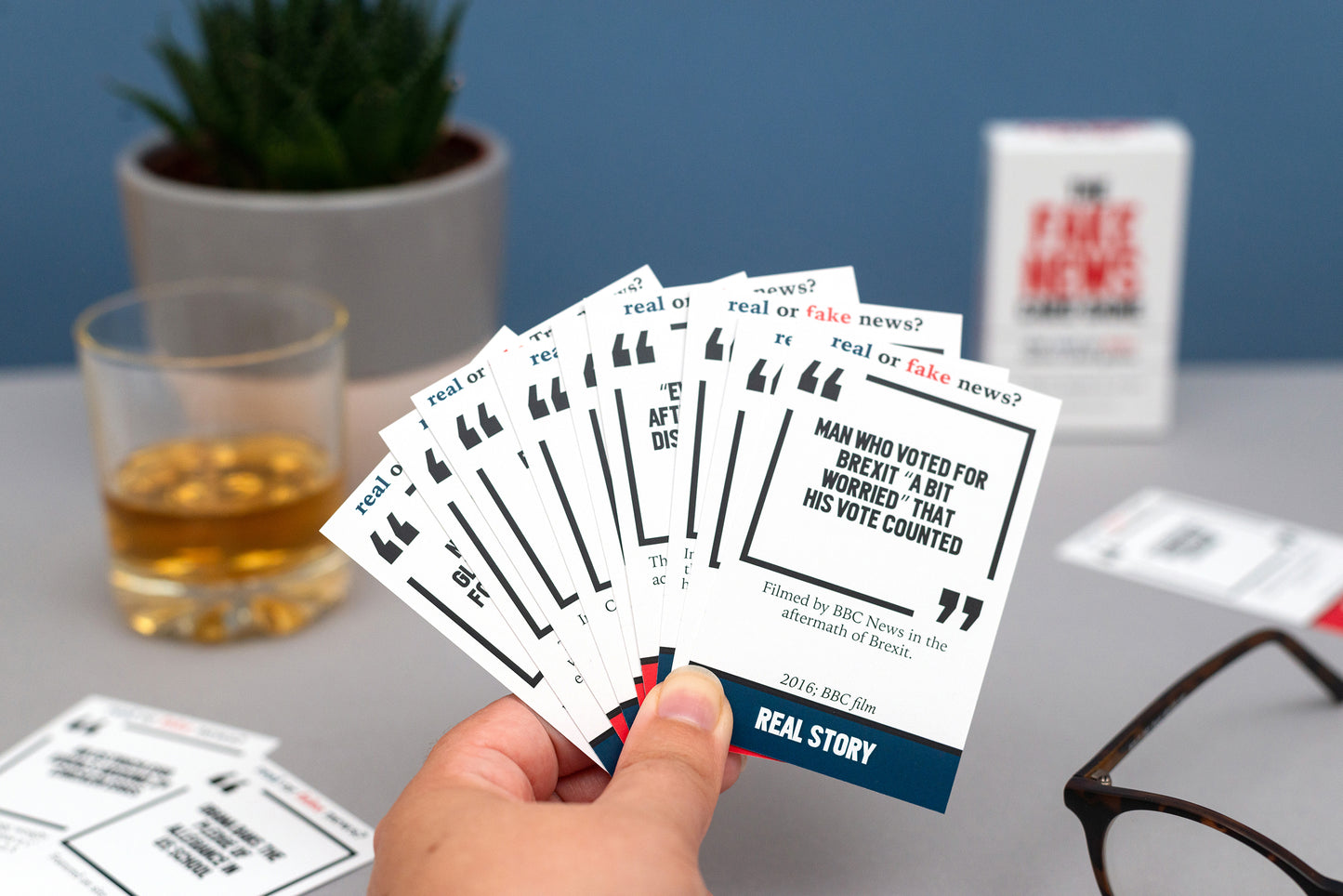 Hand holding a set of 'Real or Fake News?' cards with a glass of whisky and plant in the background.