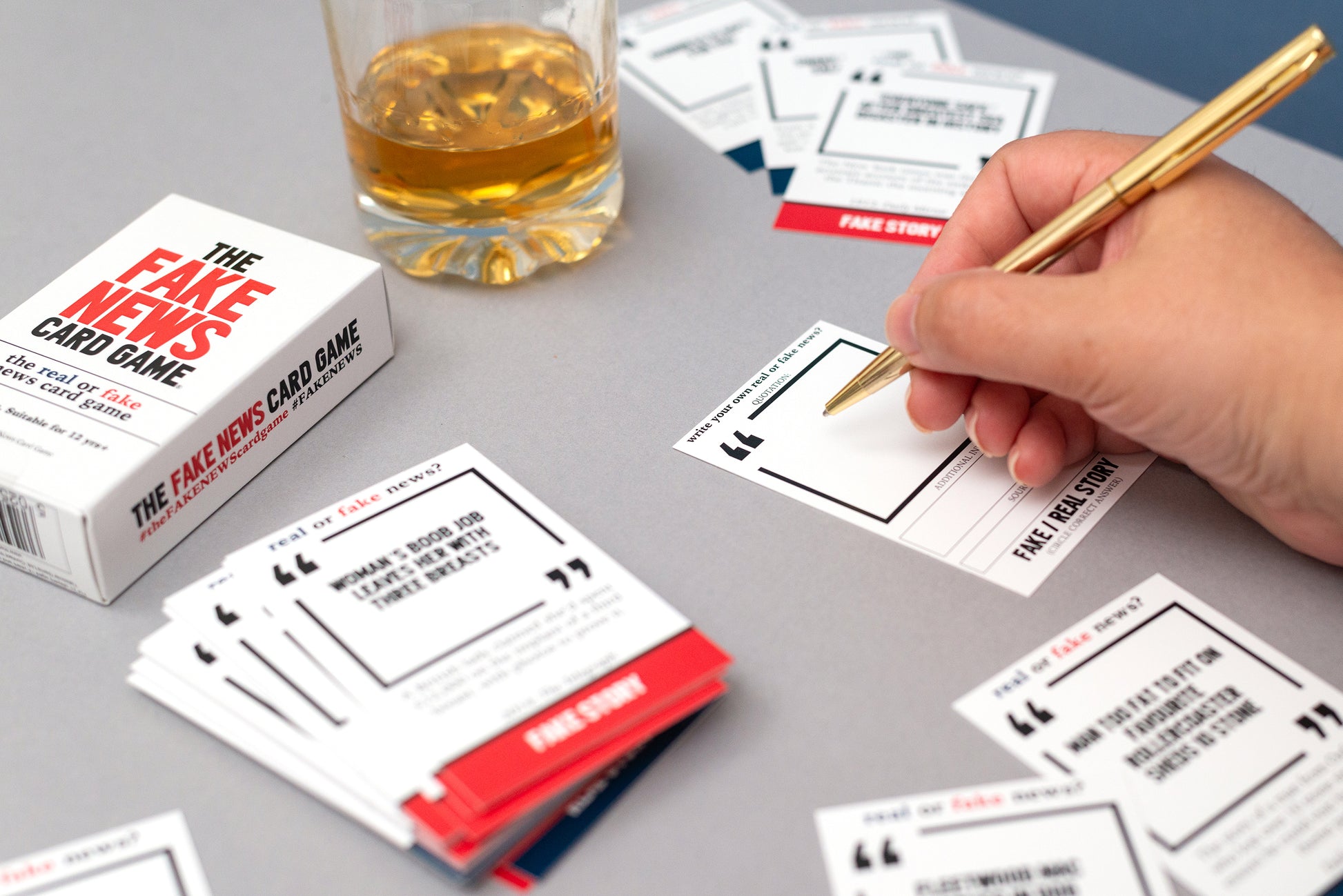 The Fake News Card Game with cards and a glass of whisky on a table.