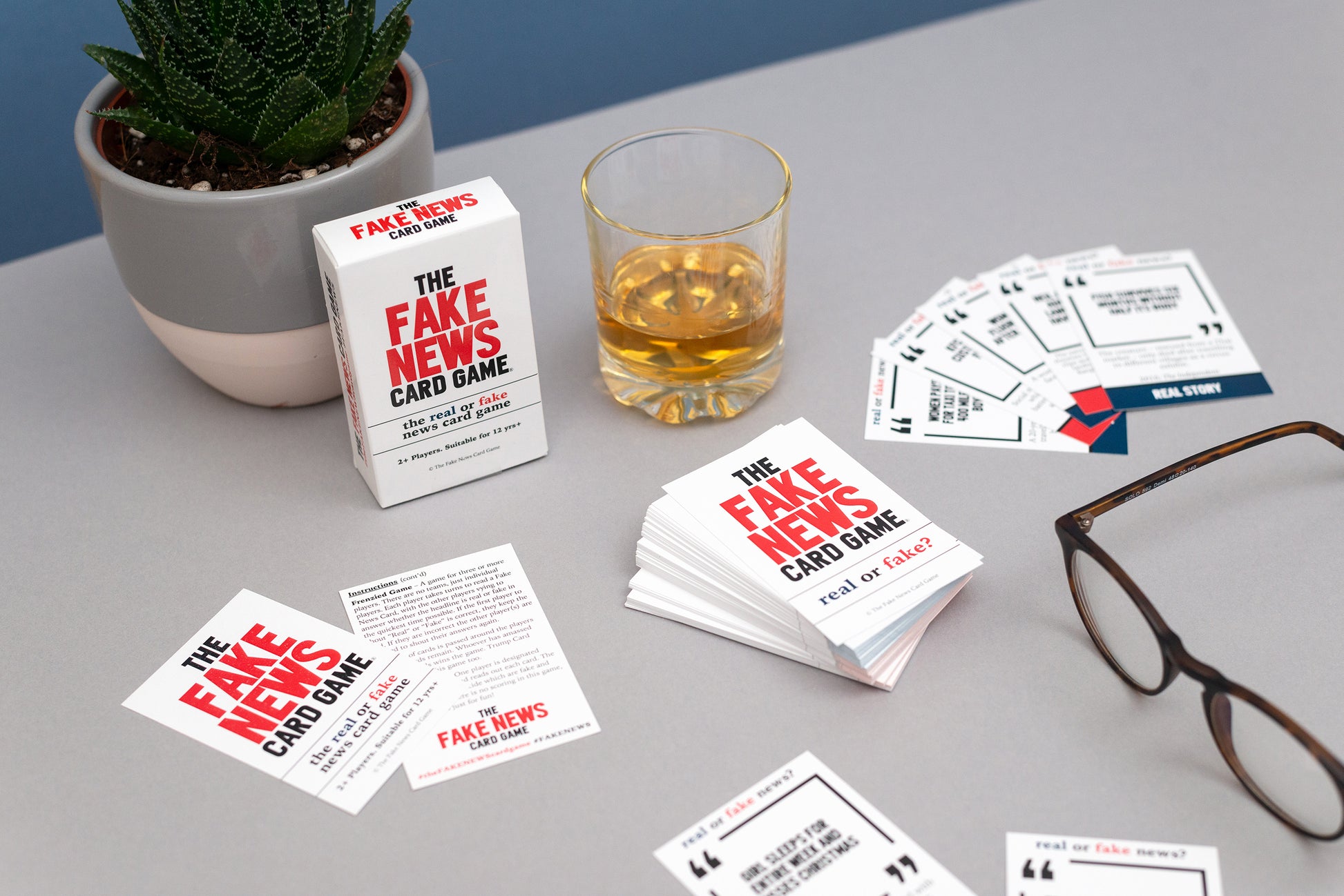 The Fake News card game with cards, packaging, and a glass of whisky on a table.