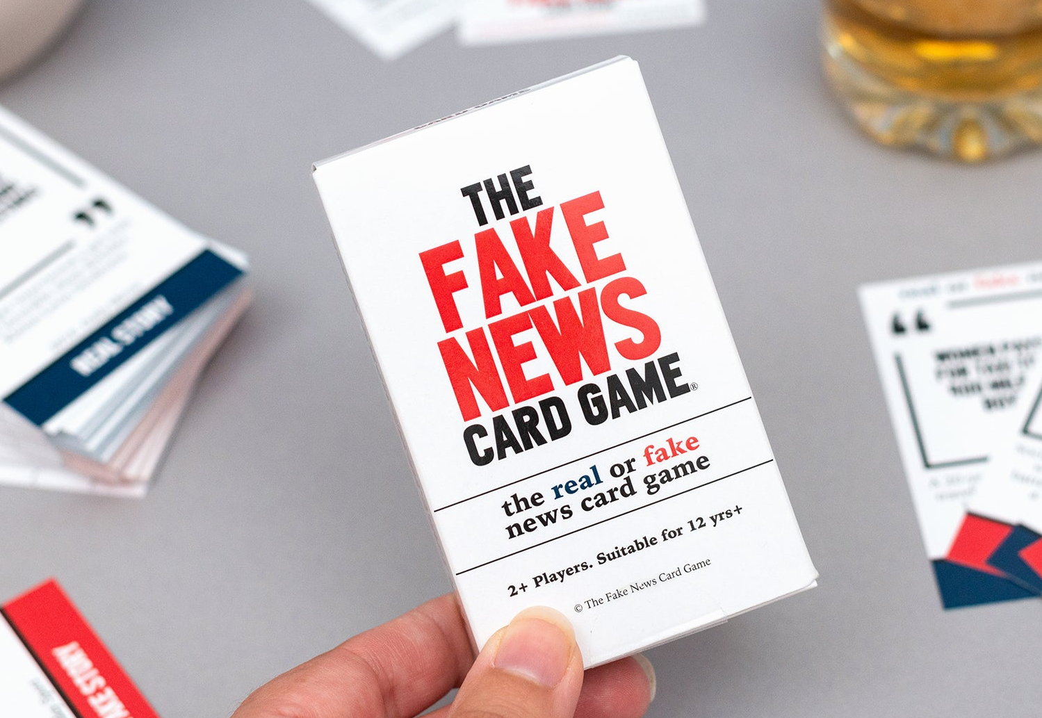Card game packaging titled 'The Fake News Card Game' held by a hand on a gray surface with additional cards.