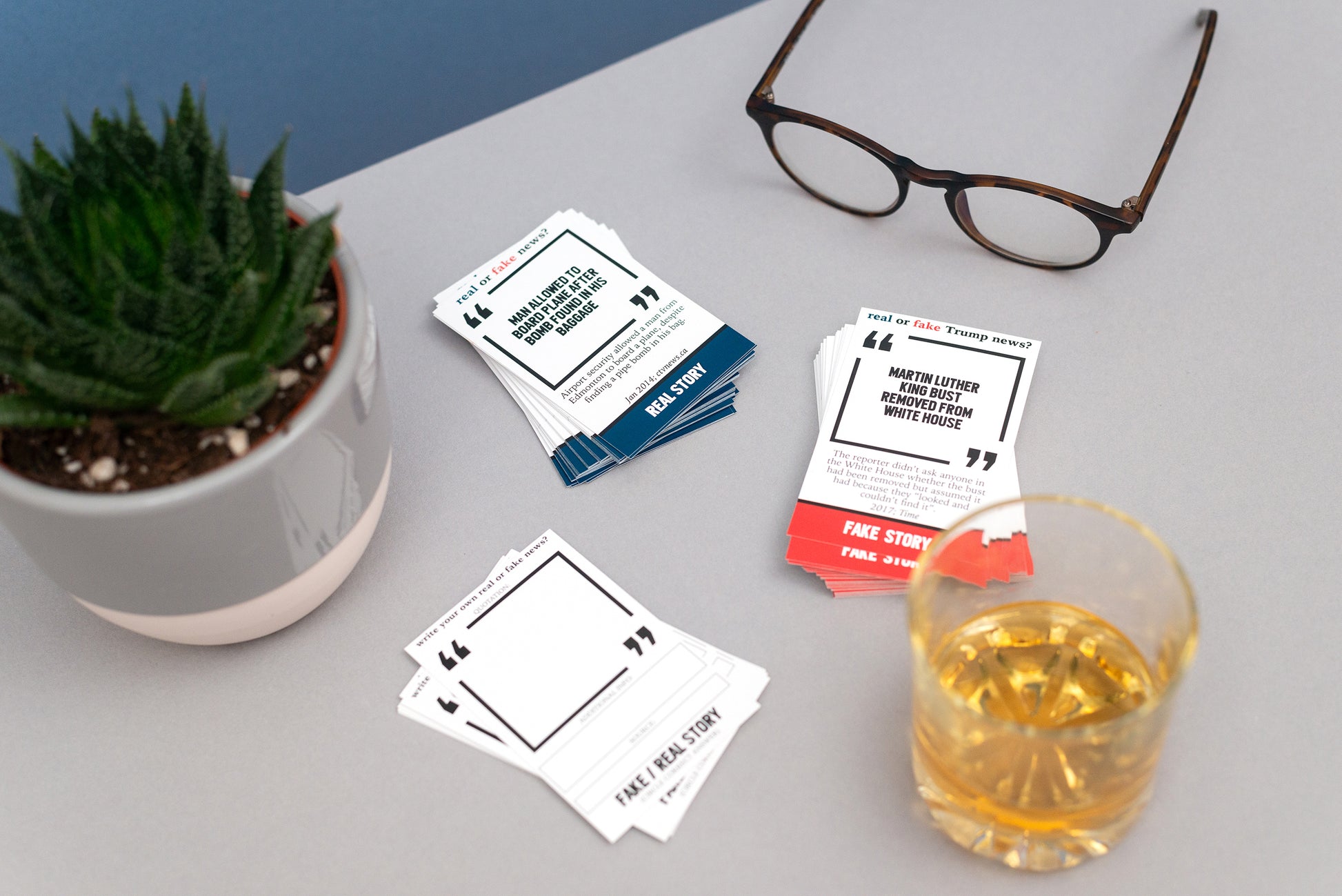 Playing cards with text on a table with a plant, glasses, and a glass of whisky.