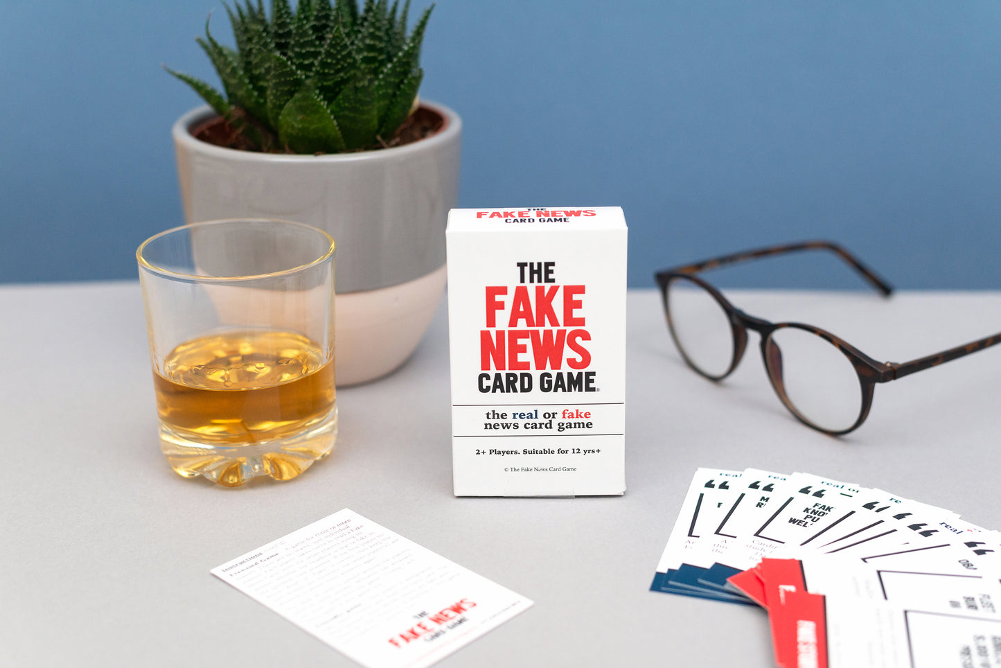 The Fake News Card Game with cards, a glass of whisky, and a plant on a table.